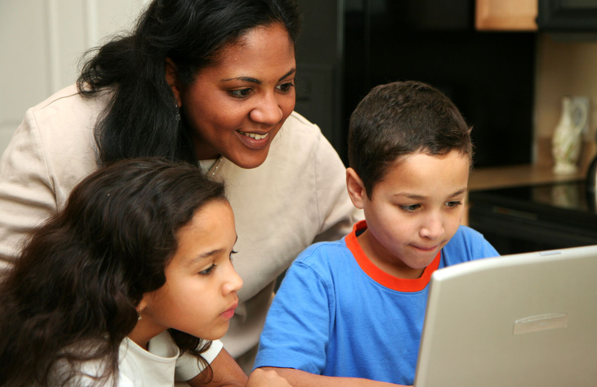A mother with a young girl and boy use a laptop at home.